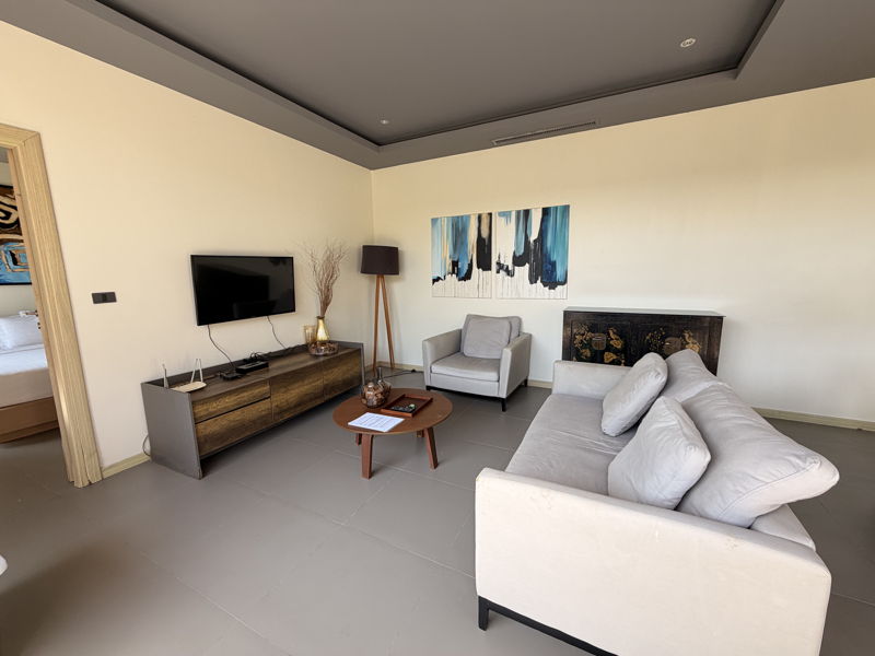 Modern living room with a wall-mounted TV on a wooden console, beige sofa, and light gray armchair around a round coffee table.