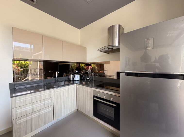 Modern L-shaped kitchen with light wood cabinets, dark countertops, and a stainless steel refrigerator and oven suite