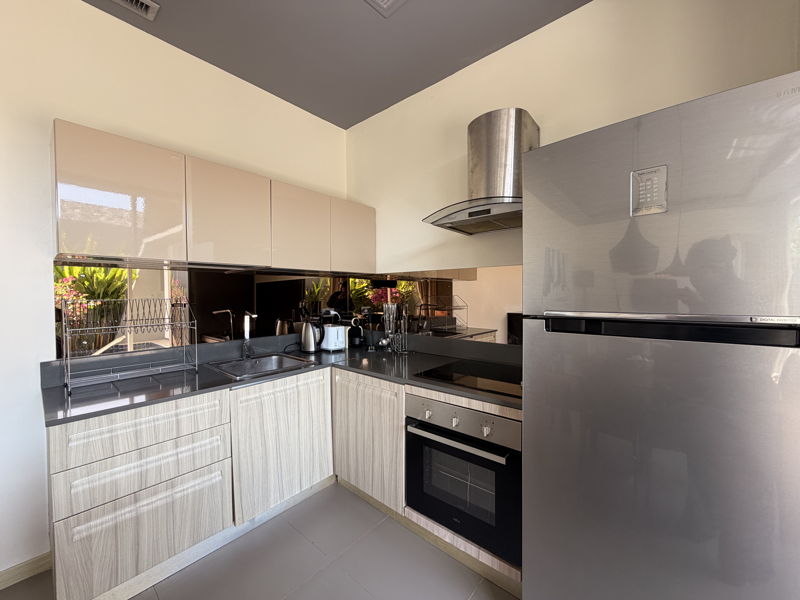 Modern L-shaped kitchen with light wood cabinets, dark countertops, and a stainless steel refrigerator and oven suite