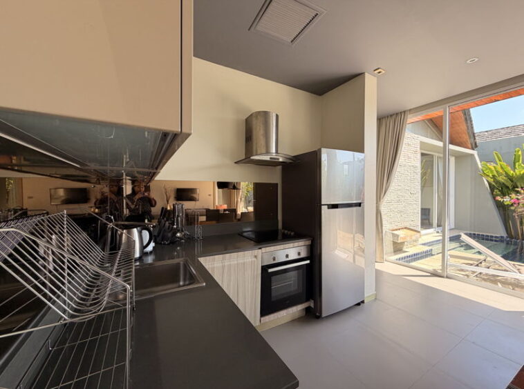 Modern kitchen with stainless-steel appliances, black countertops, a dish rack, and a sunlit pool area visible through sliding glass doors.