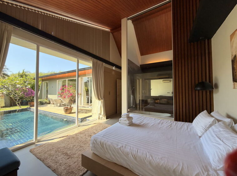 Bright bedroom with a king bed in white linens facing a large glass wall that opens to a poolside patio.