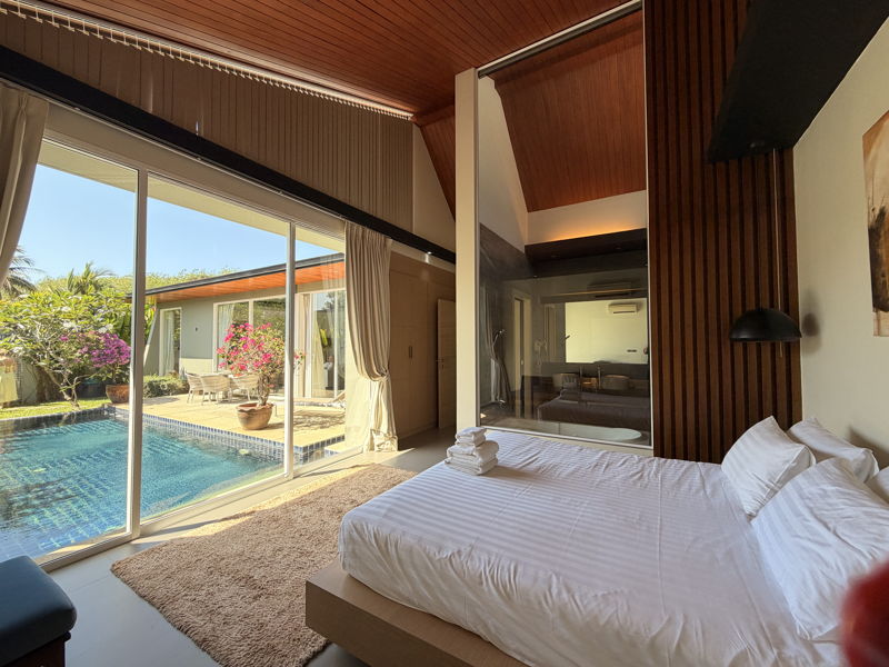 Bright bedroom with a king bed in white linens facing a large glass wall that opens to a poolside patio.