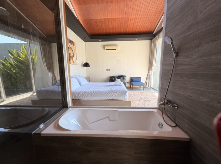 Open-plan hotel bathroom connected to a bedroom, featuring a white soaking tub, glass wall, and dark wood surroundings.