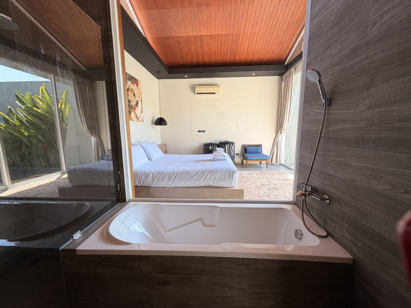 Open-plan hotel bathroom connected to a bedroom, featuring a white soaking tub, glass wall, and dark wood surroundings.