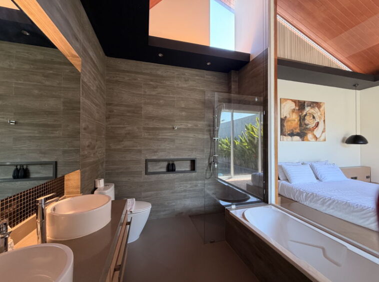 Modern en-suite bath with dual sinks, toilet, glass shower, and a bathtub beside a large glass wall facing a bedroom view?urban rustic style with gray tile walls.