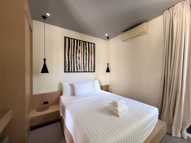 Modern hotel room with a neatly made bed in white linens, two black pendant lights, abstract wall art, and stacked towels on the foot of the bed.