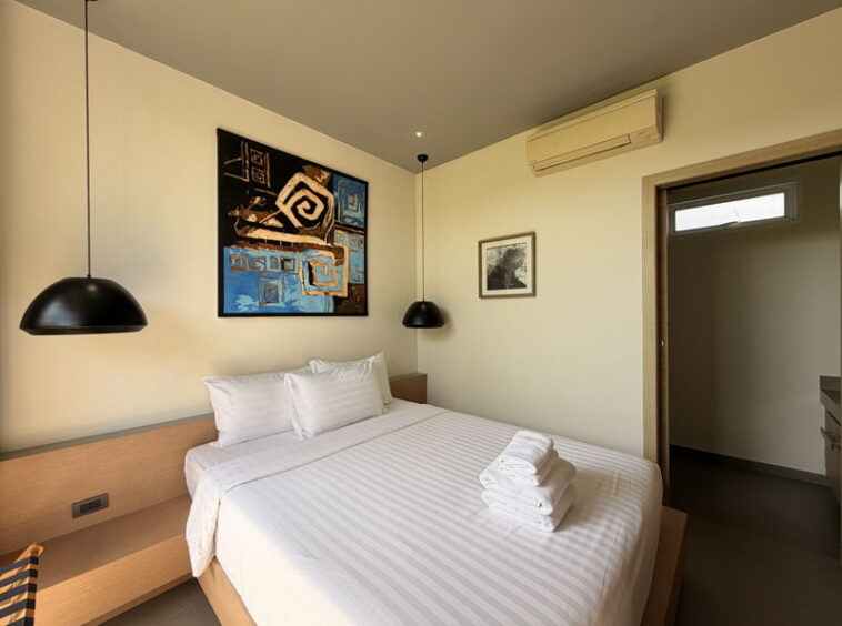 Double bed in a minimalist hotel room with white linens, stacked towels, and black pendant lamps on both sides of the headboard.