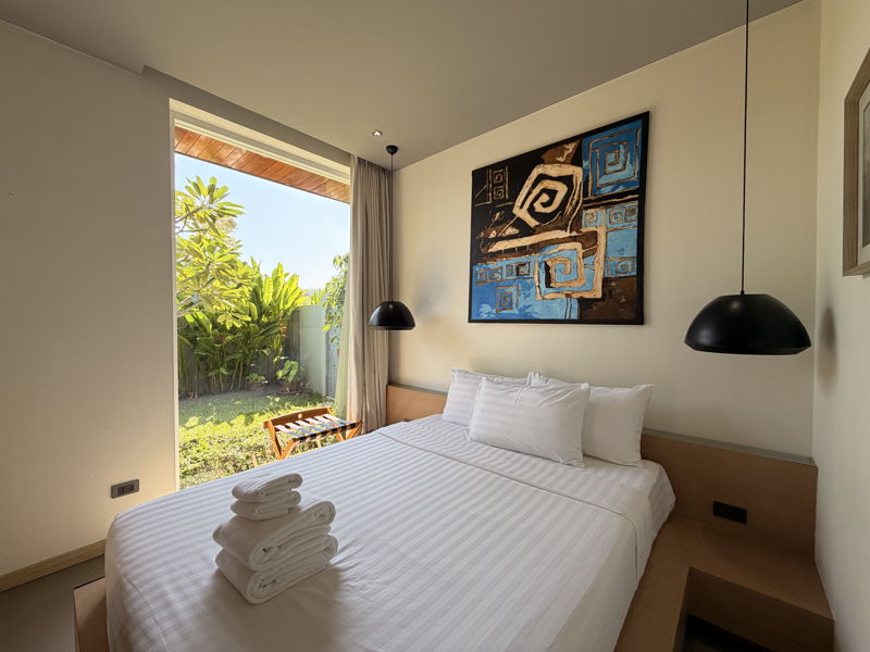 Hotel bedroom with white bedding and folded towels, black pendant lamps, abstract blue artwork, and a glass door opening to a sunny garden.