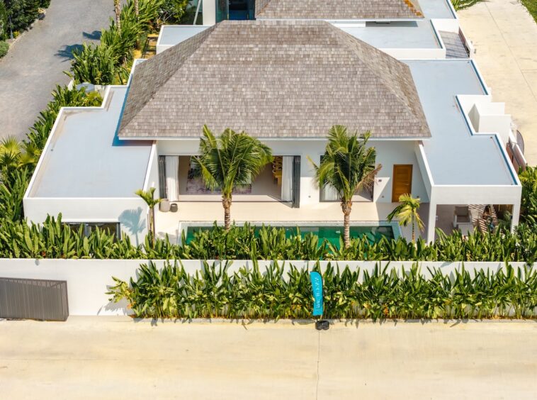 Aerial view of a modern white villa with a thatched roof, palm trees, and a rectangular pool amid tropical landscaping along a street.