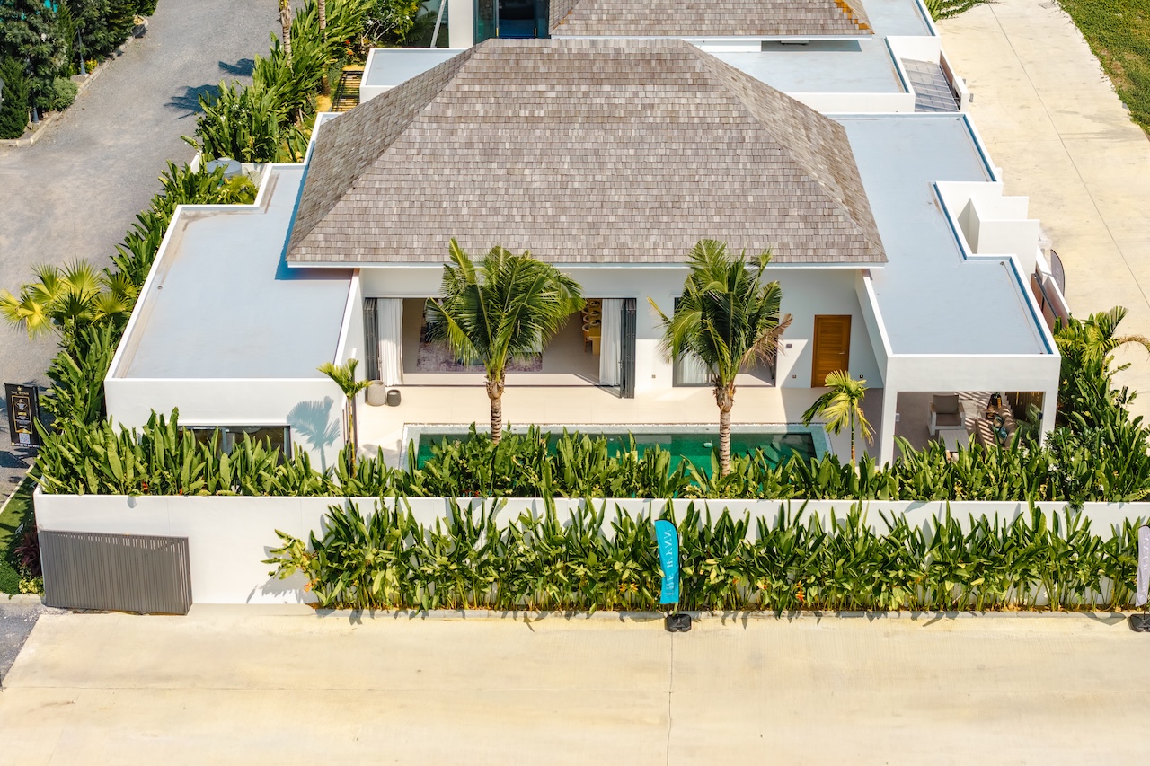 Aerial view of a modern white villa with a thatched roof, palm trees, and a rectangular pool amid tropical landscaping along a street.