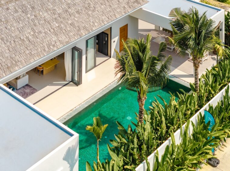 Aerial view of a modern villa with a thatched roof, glass doors open to a shaded living area, and a turquoise rectangular pool flanked by tropical plants and palm trees.