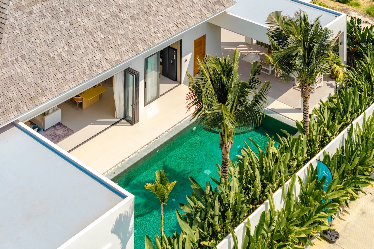 Aerial view of a modern villa with a thatched roof, glass doors open to a shaded living area, and a turquoise rectangular pool flanked by tropical plants and palm trees.