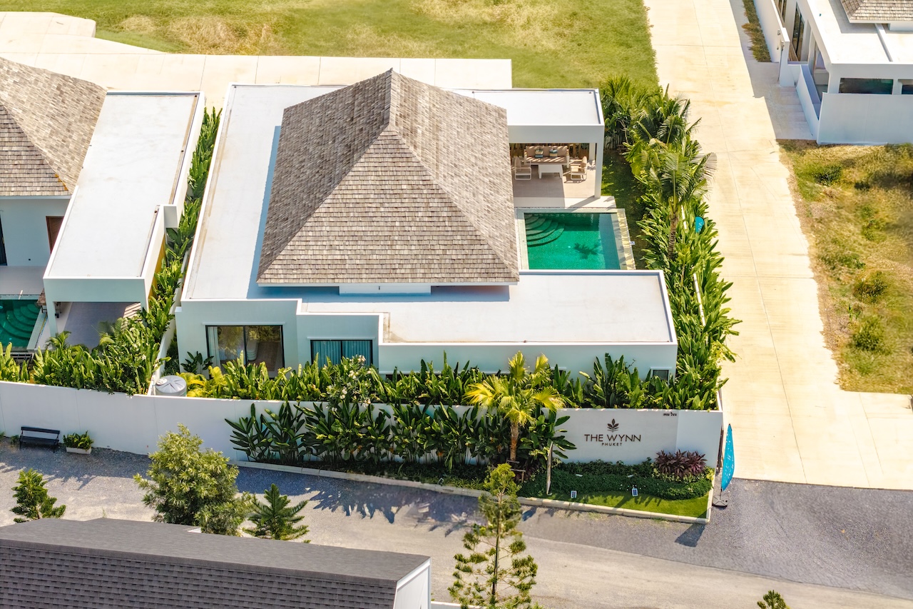 Aerial view of a modern villa with a central pyramid-shaped thatched roof, a rectangular pool, and tropical landscaping around The Wynn Phuket.