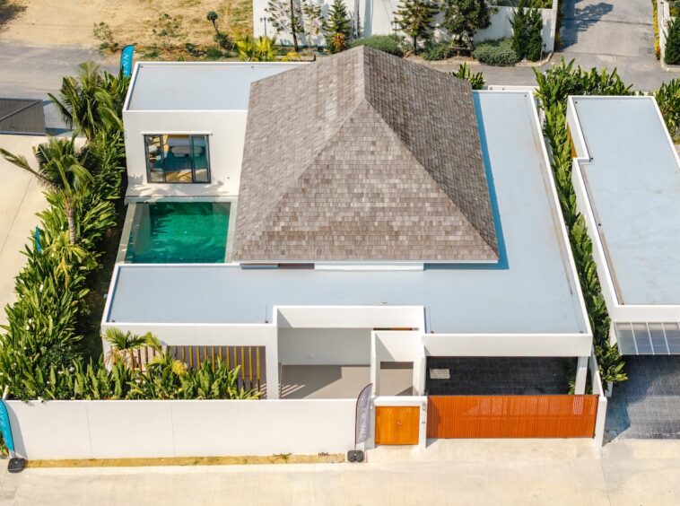 Aerial view of a modern white house with a rectangular turquoise pool, surrounded by tropical plants and an orange front gate.