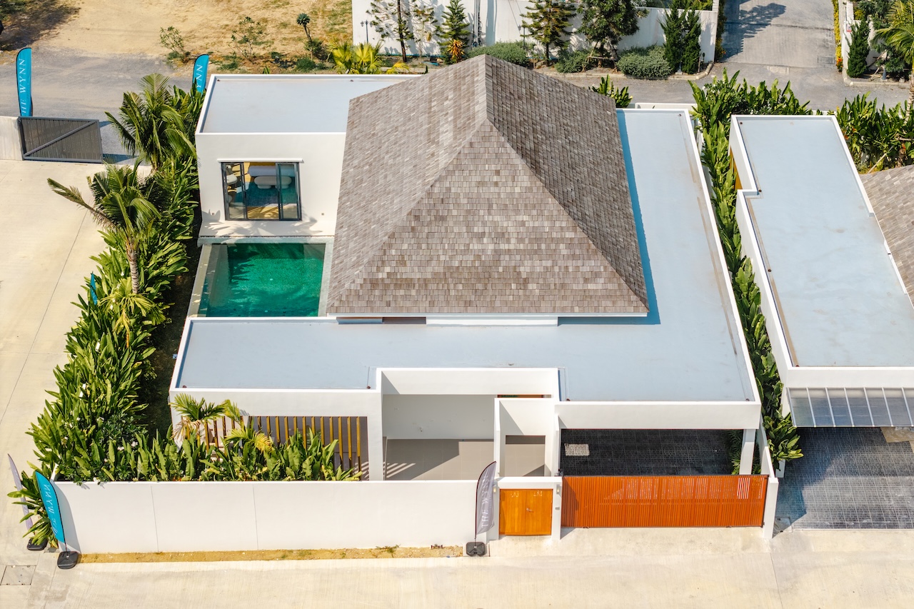 Aerial view of a modern white house with a rectangular turquoise pool, surrounded by tropical plants and an orange front gate.