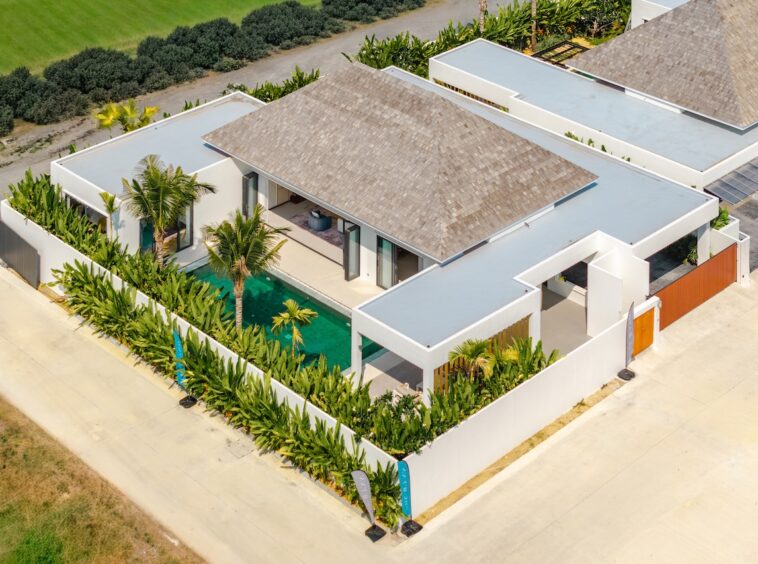 Aerial view of a modern white villa with a rectangular pool, palm trees, and a gated garden courtyard surrounded by greenery.