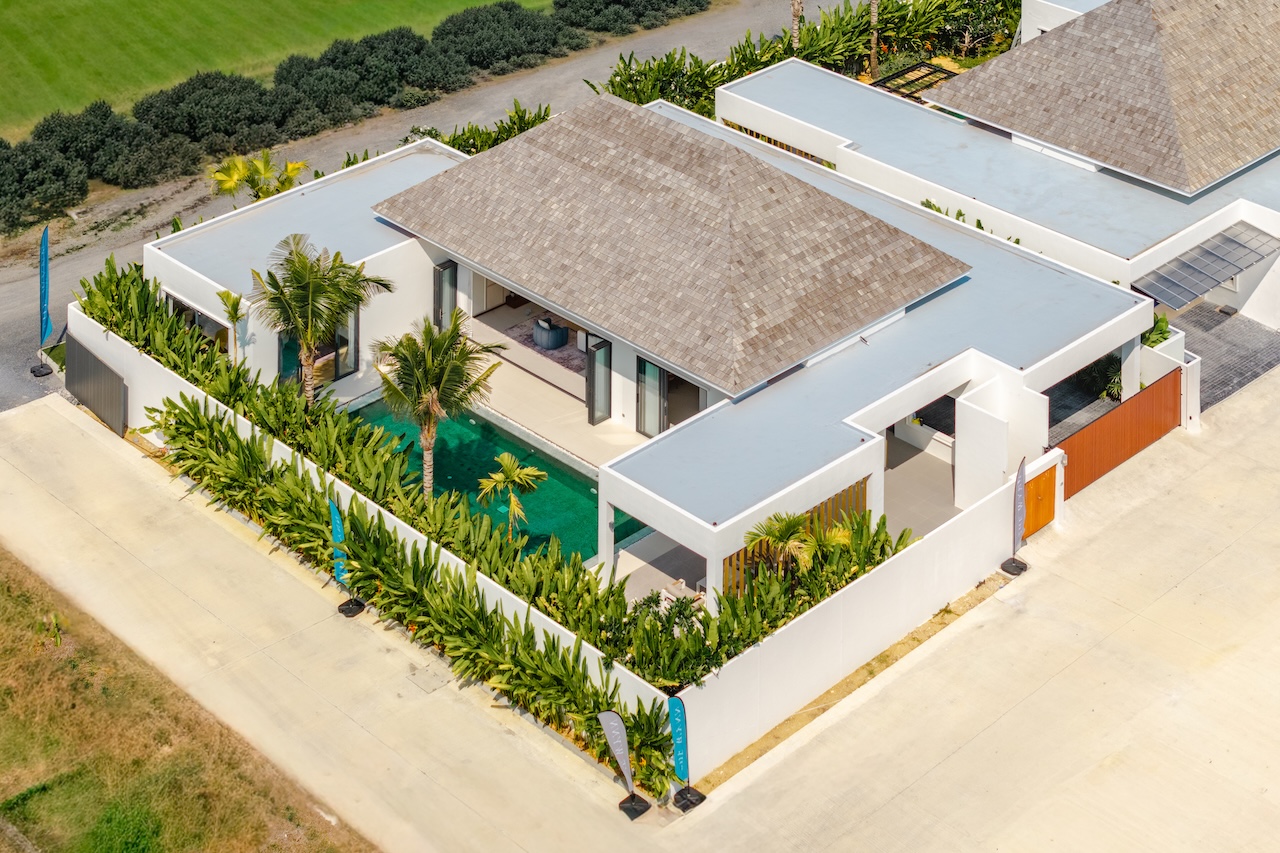 Aerial view of a modern white villa with a rectangular pool, palm trees, and a gated garden courtyard surrounded by greenery.
