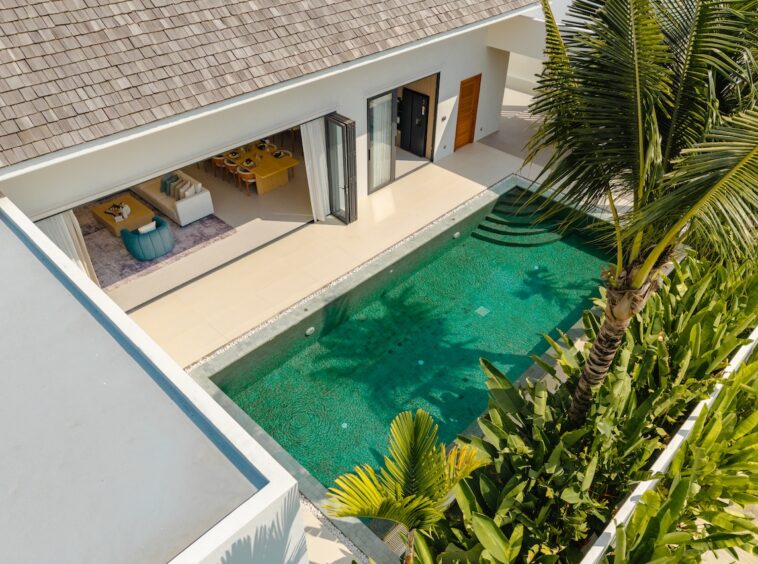 Aerial view of a modern villa with a turquoise rectangular pool, open living area, and tropical plants nearby.
