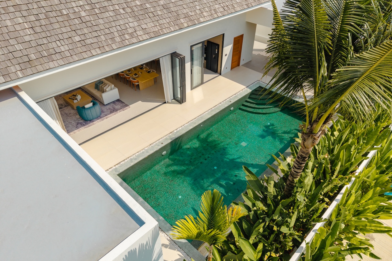 Aerial view of a modern villa with a turquoise rectangular pool, open living area, and tropical plants nearby.