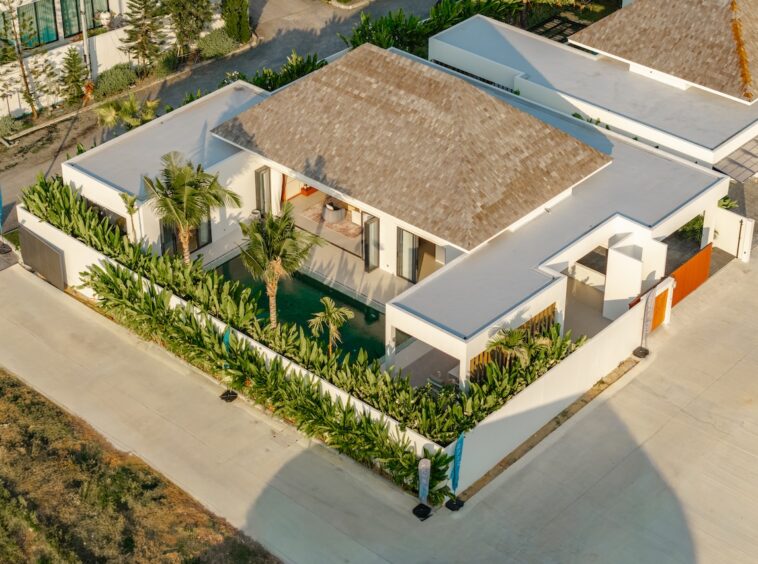 Aerial view of a modern white villa with a brown roof, glass doors, and a rectangular pool bordered by tropical plants.