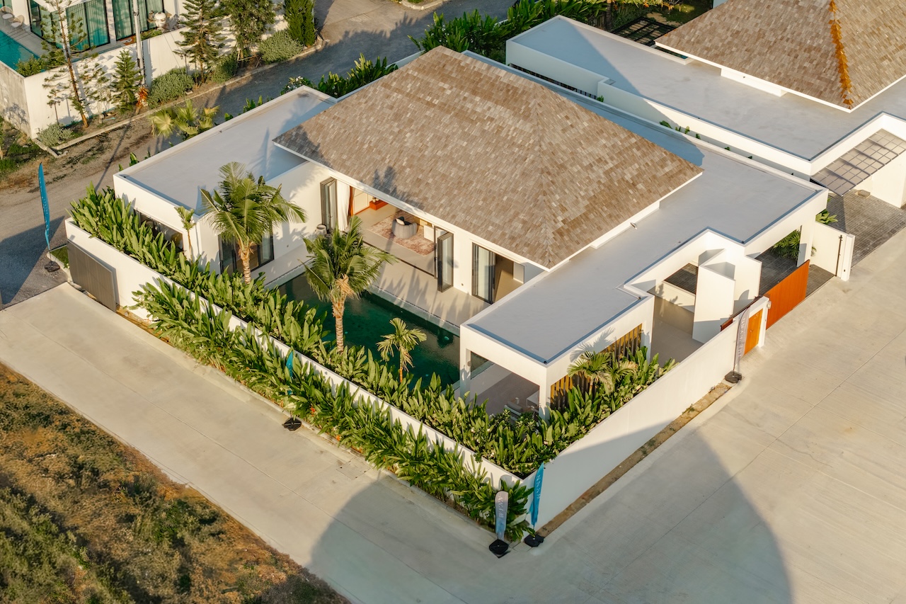Aerial view of a modern white villa with a brown roof, glass doors, and a rectangular pool bordered by tropical plants.
