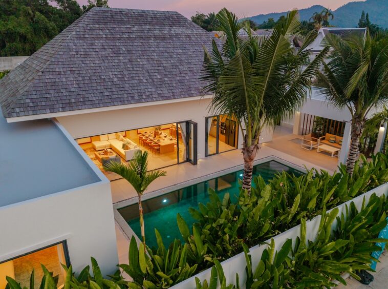 Aerial view of a modern villa with a rectangular pool, palm trees, and a sunlit open-plan living and dining area visible through glass doors.