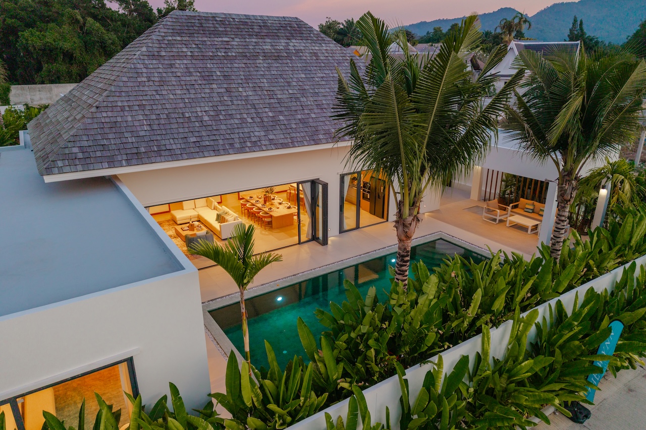 Aerial view of a modern villa with a rectangular pool, palm trees, and a sunlit open-plan living and dining area visible through glass doors.