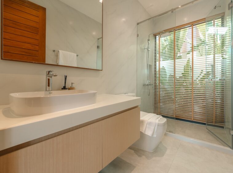 Modern bathroom with a white vessel sink on a light wooden vanity, large mirror, and glass-enclosed shower with wooden blinds outside.