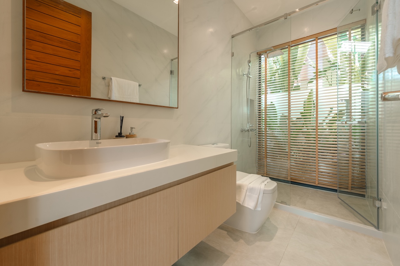 Modern bathroom with a white vessel sink on a light wooden vanity, large mirror, and glass-enclosed shower with wooden blinds outside.