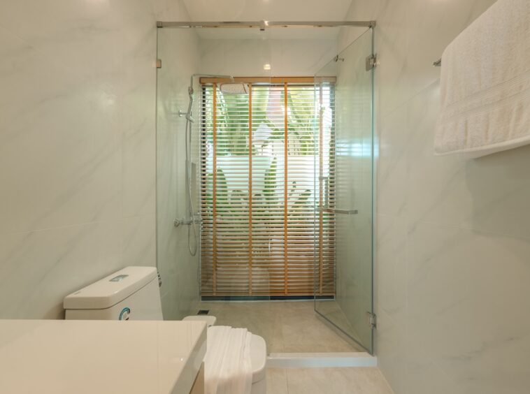 Modern bathroom with glass-enclosed shower, white toilet, and towel rack; window with wooden blinds and greenery outside