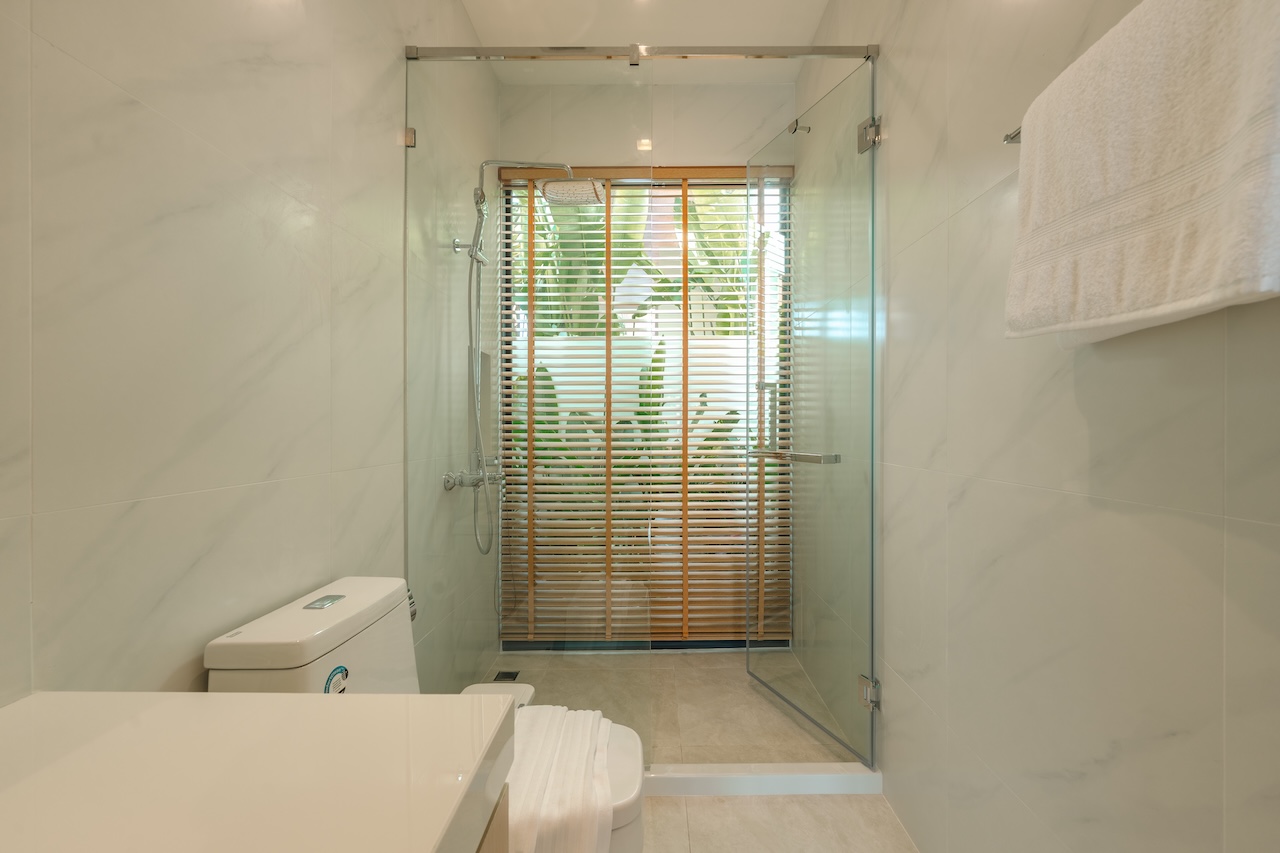 Modern bathroom with glass-enclosed shower, white toilet, and towel rack; window with wooden blinds and greenery outside