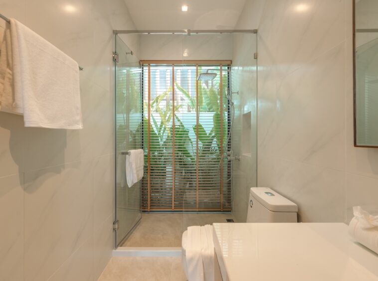Bright modern bathroom with glass shower enclosure, white marble walls, a toilet, and a white countertop; wooden blinds and greenery outside.
