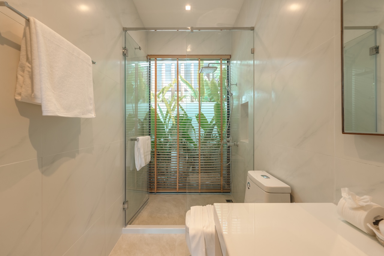 Bright modern bathroom with glass shower enclosure, white marble walls, a toilet, and a white countertop; wooden blinds and greenery outside.