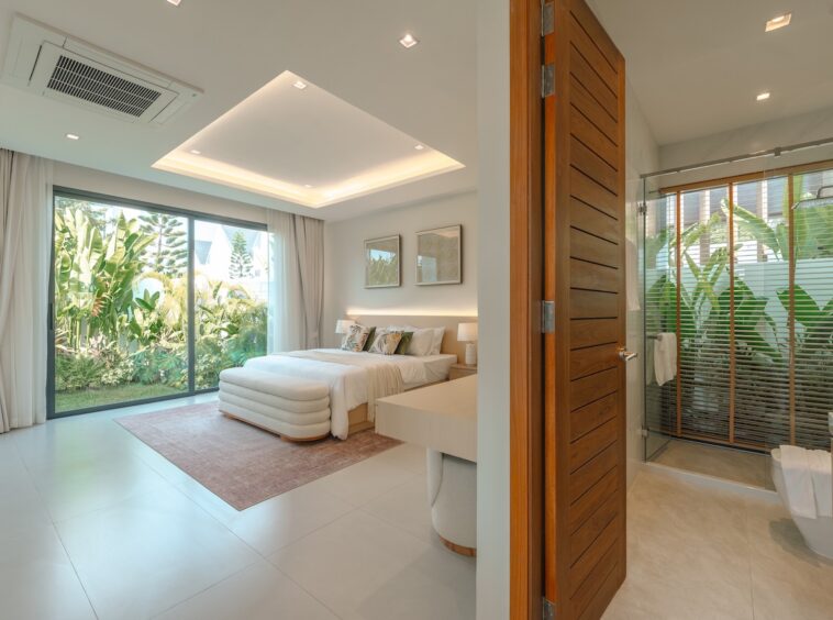 Bright hotel room with a king bed, white bedding, and a plush bench at the foot, opening to a lush garden through large sliding glass doors.