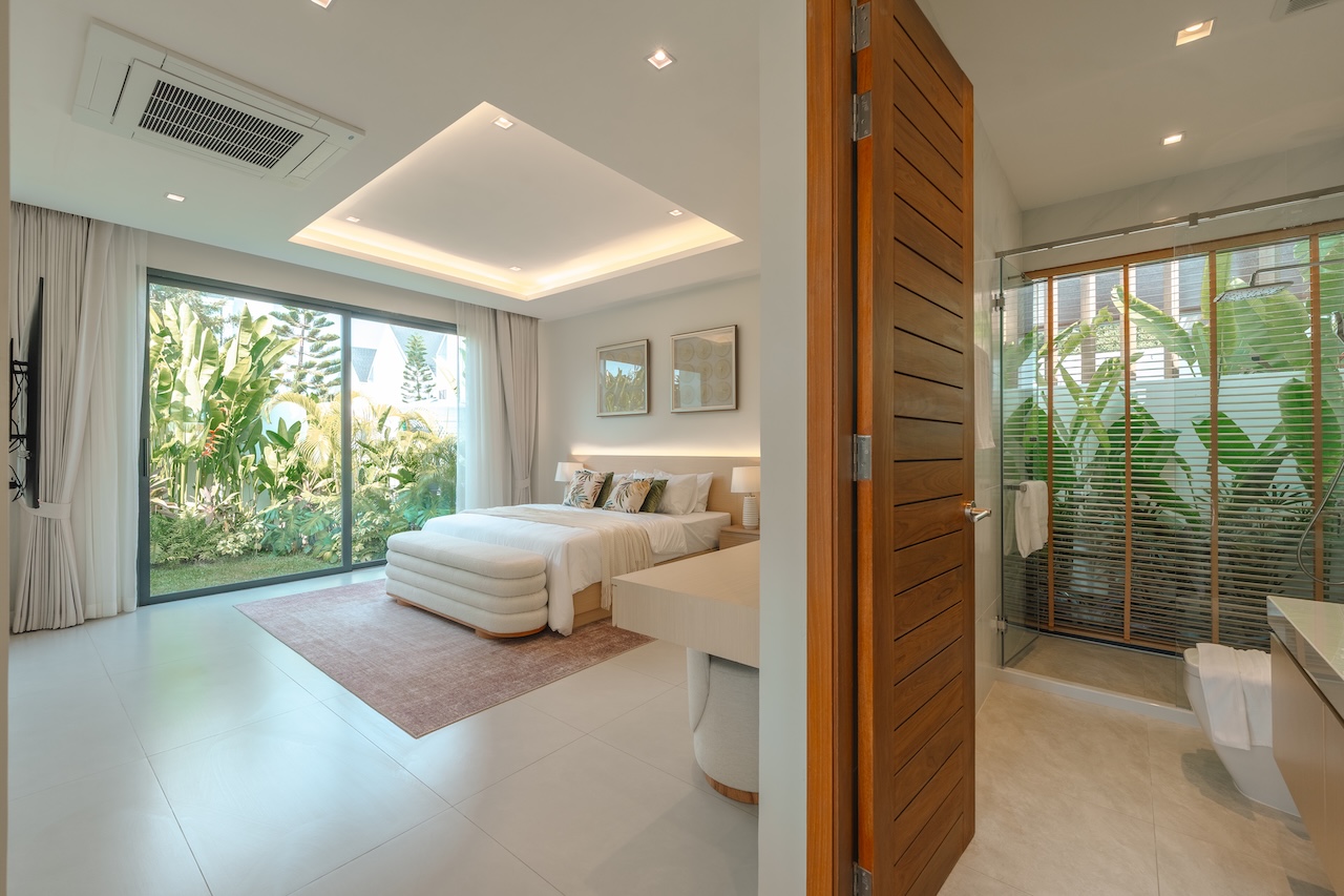 Bright hotel room with a king bed, white bedding, and a plush bench at the foot, opening to a lush garden through large sliding glass doors.