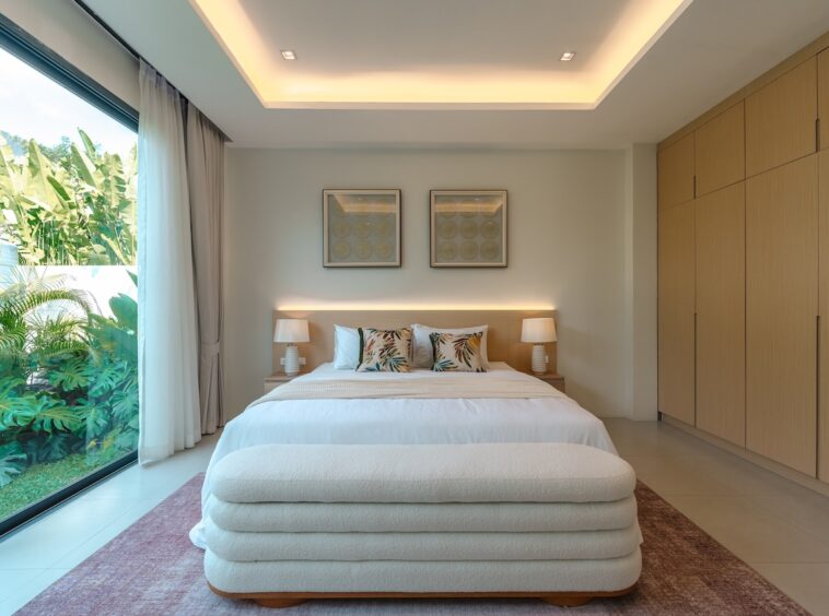 Modern bedroom with a large glass wall showing tropical plants outside, and a white bed with botanical cushions.