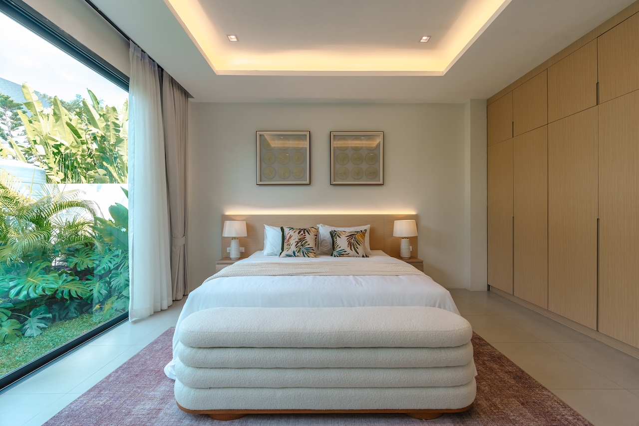 Modern bedroom with a large glass wall showing tropical plants outside, and a white bed with botanical cushions.