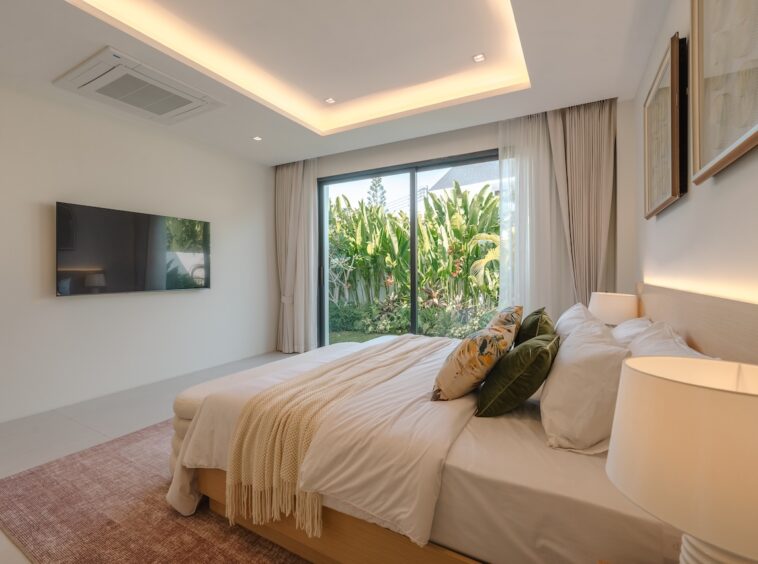 Modern hotel bedroom with a king bed, neutral linens, and a glass wall opening to a tropical garden, bright and inviting.