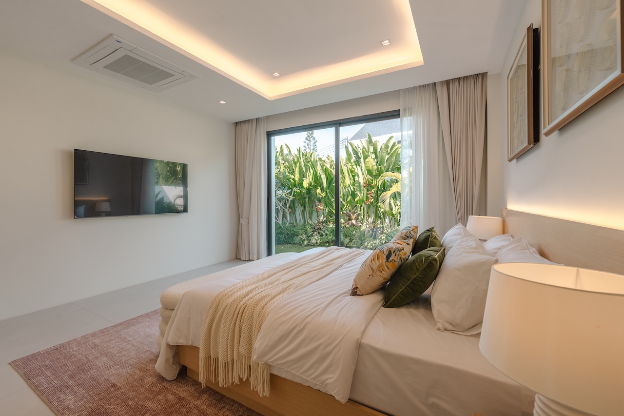 Modern hotel bedroom with a king bed, neutral linens, and a glass wall opening to a tropical garden, bright and inviting.