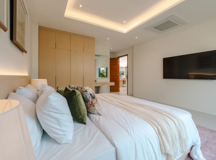 Bright modern bedroom with a low wood headboard, white bedding, green and floral cushions, and a wall-mounted TV.