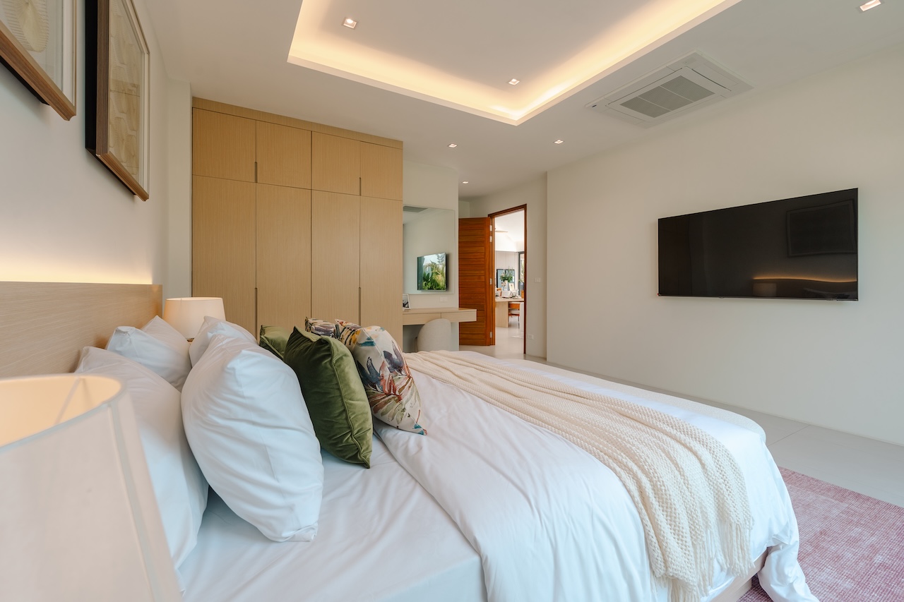 Bright modern bedroom with a low wood headboard, white bedding, green and floral cushions, and a wall-mounted TV.
