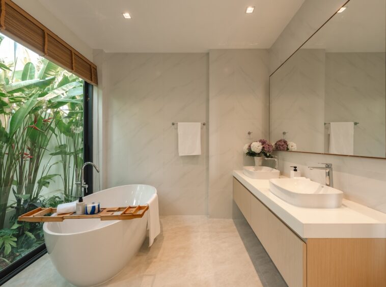 Freestanding white tub by a large glass wall with tropical plants, wooden bath tray, and a double sink vanity with a large mirror.