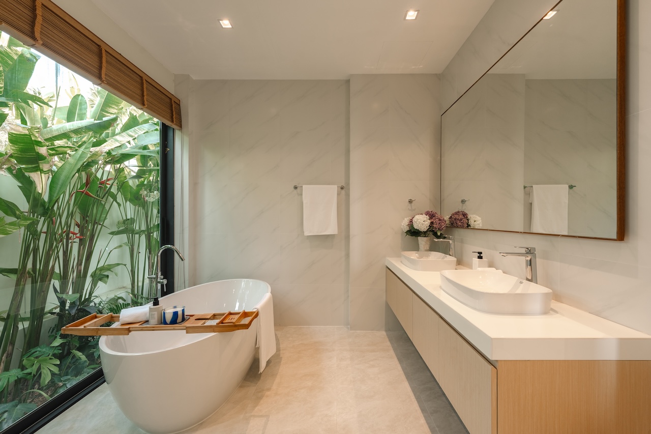 Freestanding white tub by a large glass wall with tropical plants, wooden bath tray, and a double sink vanity with a large mirror.