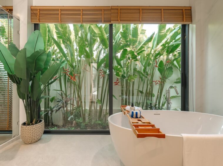 Freestanding white bathtub with a wooden bath tray holding toiletries, in a bright bathroom overlooking tropical plants via a large glass window.