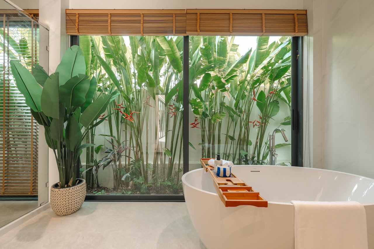 Freestanding white bathtub with a wooden bath tray holding toiletries, in a bright bathroom overlooking tropical plants via a large glass window.