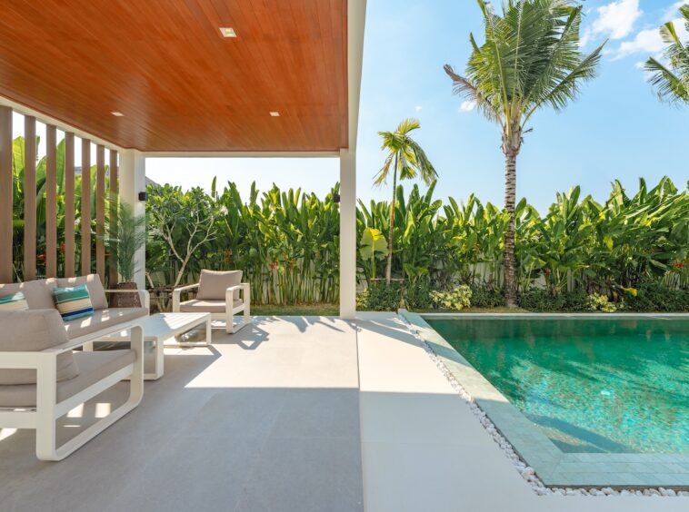 Patio with cream outdoor furniture beside a turquoise pool, surrounded by tropical plants and palm trees in bright sunshine.