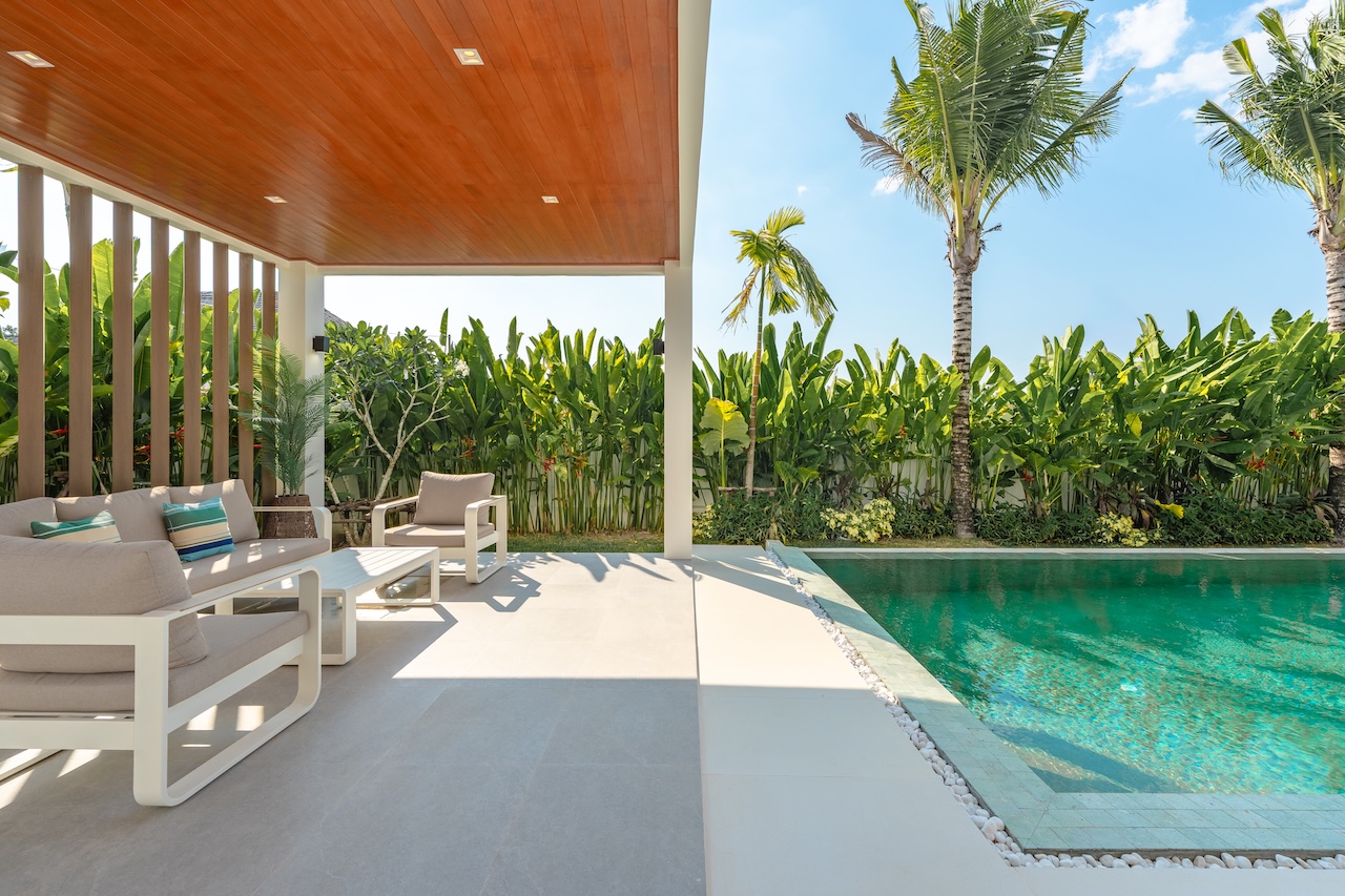 Patio with cream outdoor furniture beside a turquoise pool, surrounded by tropical plants and palm trees in bright sunshine.