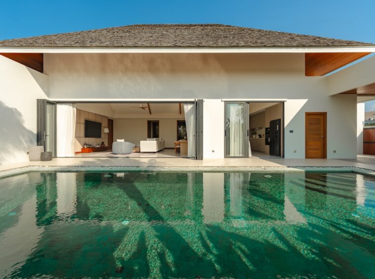 Modern house with a rectangular swimming pool in the foreground and open sliding doors leading to a bright living area.