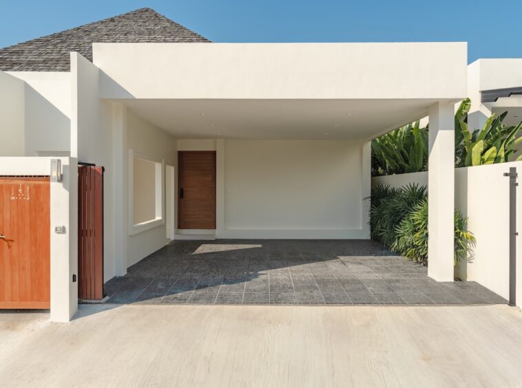 Contemporary house with a covered carport, wooden front door, and a wooden gate on the left; white walls and gray tiled driveway with tropical plants on the right.