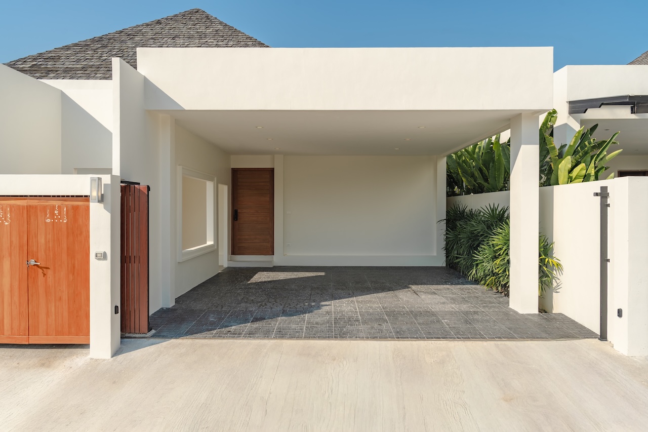 Contemporary house with a covered carport, wooden front door, and a wooden gate on the left; white walls and gray tiled driveway with tropical plants on the right.